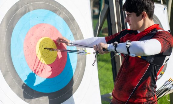 Focus on man aiming with bow and arrows at target during outdoor archery practice.